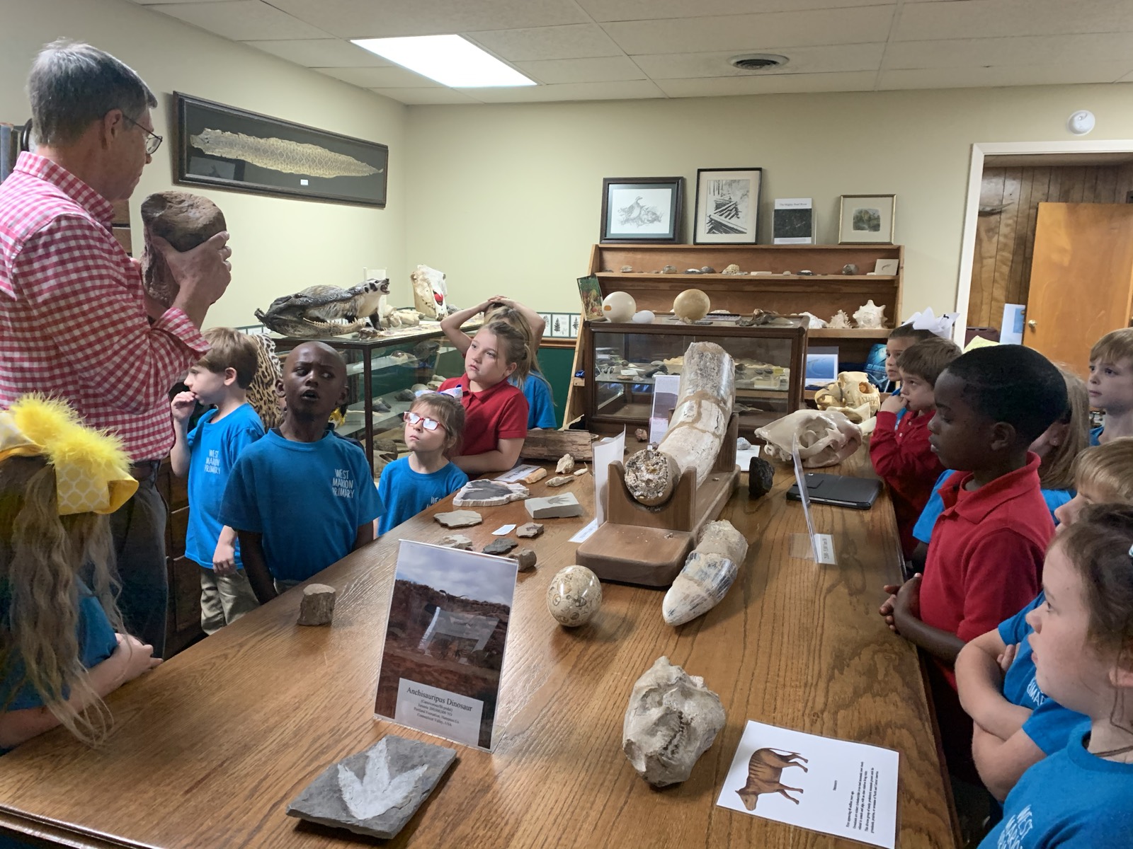Students gathered around a table of fossils, a mastodon tusk and a dinosaur track cast while Mickey Webb explains a specimen