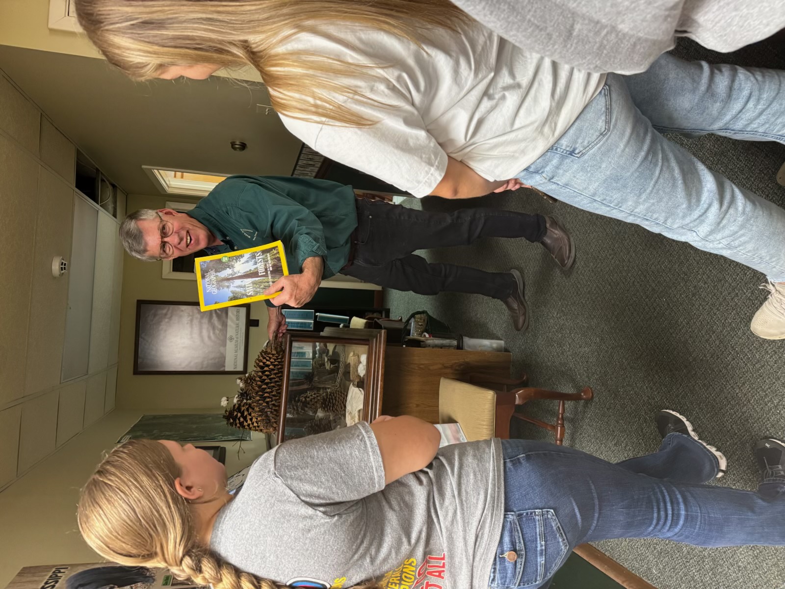 Mickey Webb showing a National Geographic issue on forests to high-school students beside a case of giant pine cones