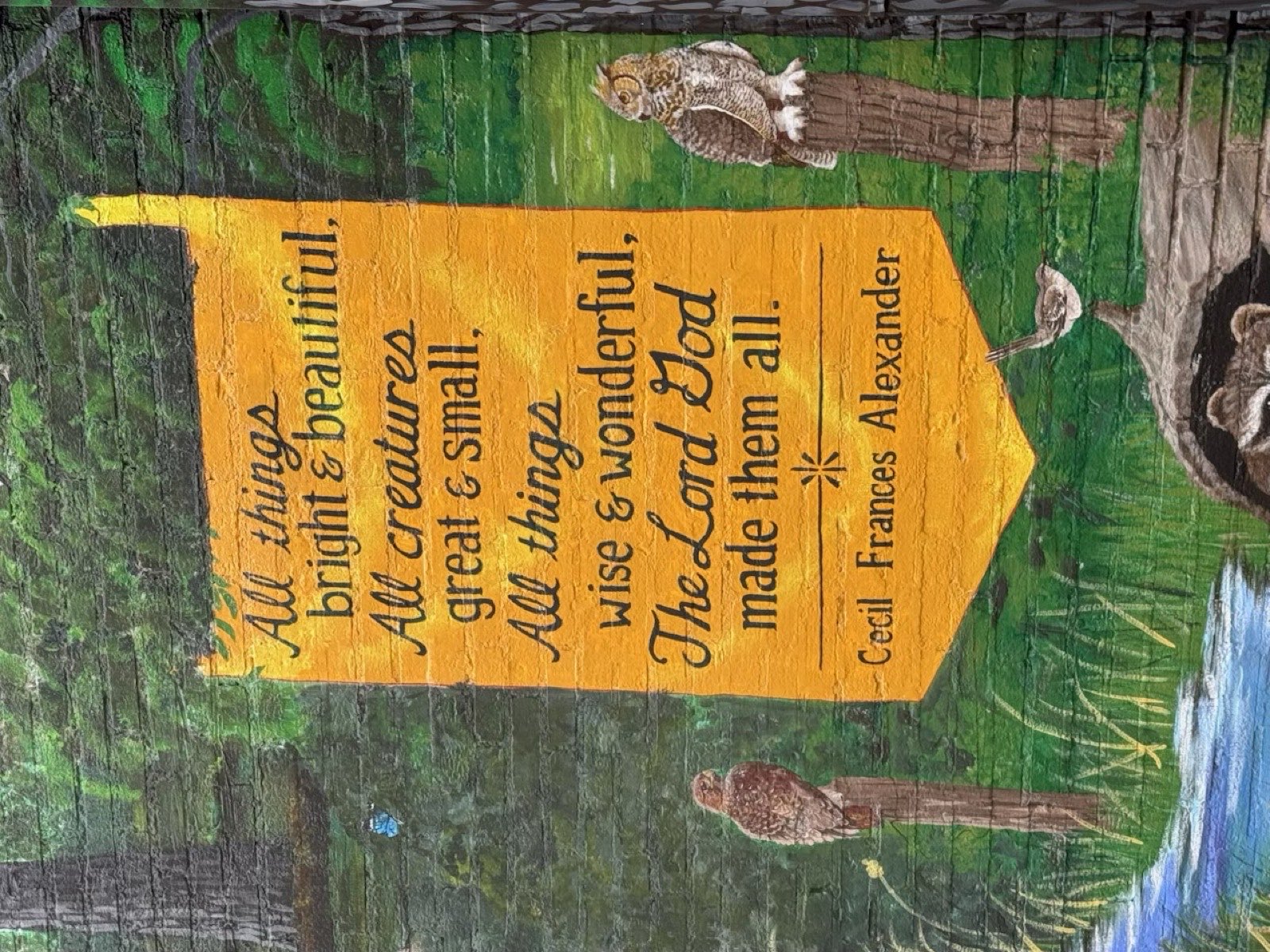 A painted banner on the museum's outdoor mural reading “All things bright and beautiful, all creatures great and small, all things wise and wonderful, the Lord God made them all” — Cecil Frances Alexander, with a hawk, an owl and a wren painted around it